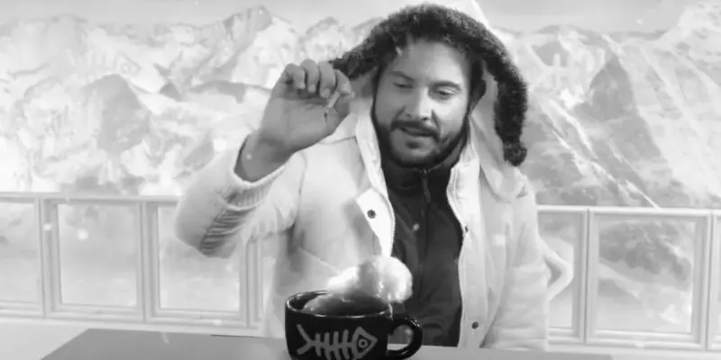 Un hombre en una montaña nevada con mucho frío, abrigo y una pequeña taza con el logo de Sy Films que empieza a arder en llamas